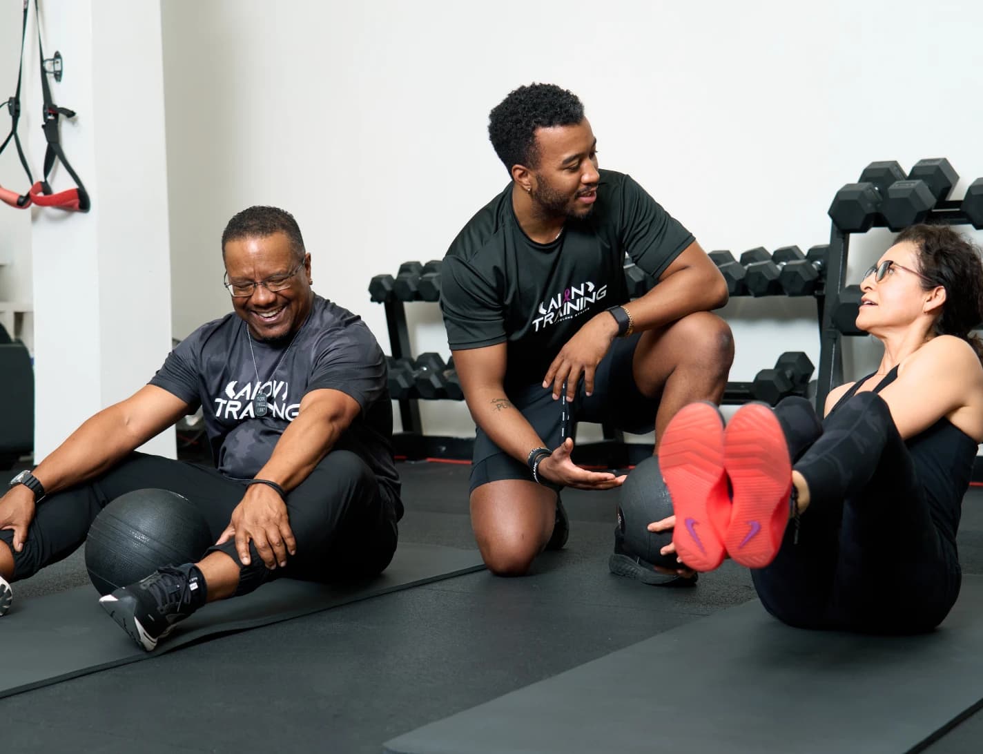 A coach sits on the floor with two members as they work through a seated core exercise.