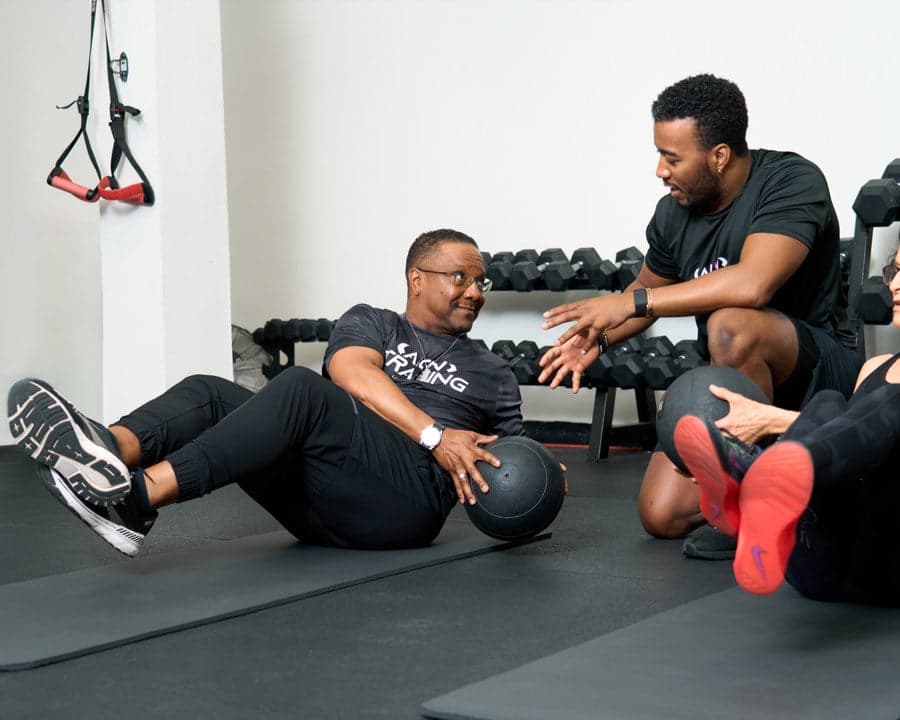 A member works through a seated medicine ball exercise on the mat while a coach guides him.