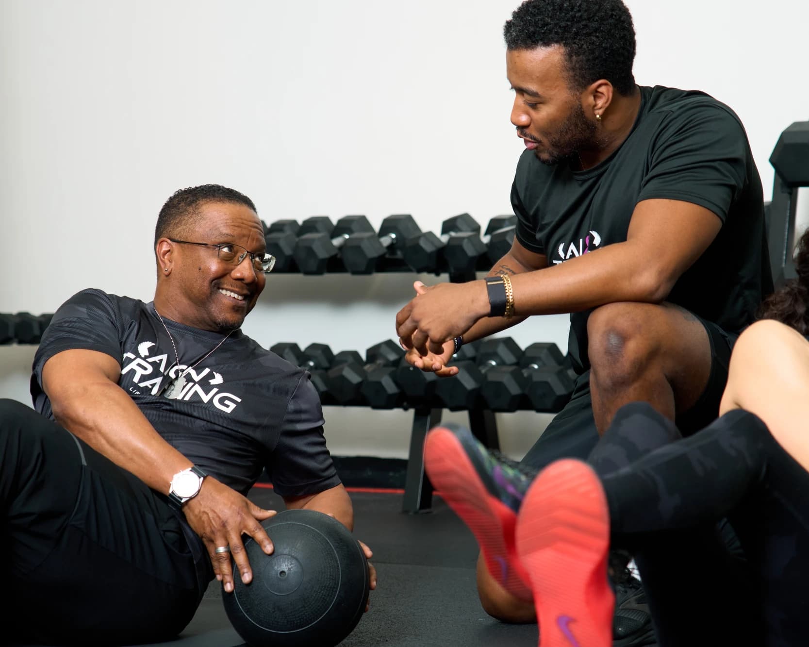 A coach kneels next to a member on the floor and coaches him through a medicine ball exercise.