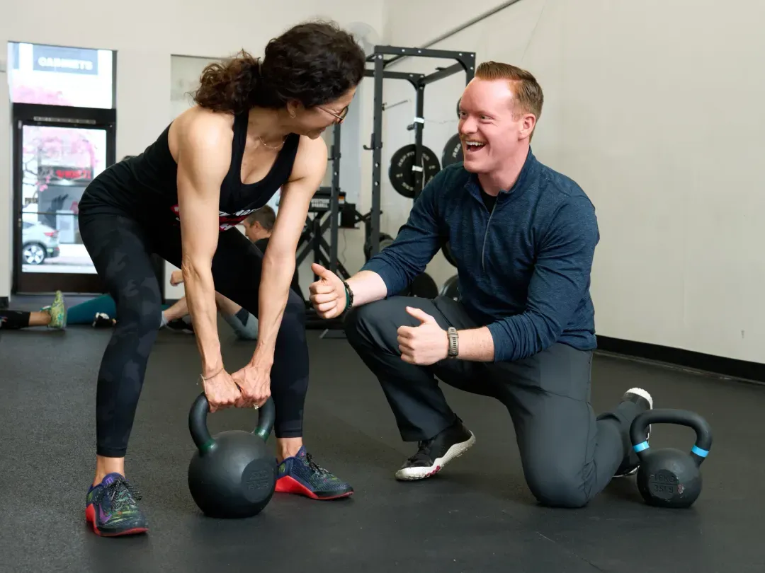A coach kneels beside a member as she lifts two kettlebells from the floor.
