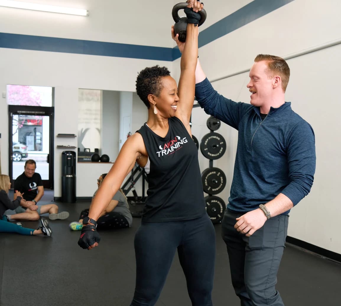 A member presses a kettlebell overhead while a coach stands alongside her.
