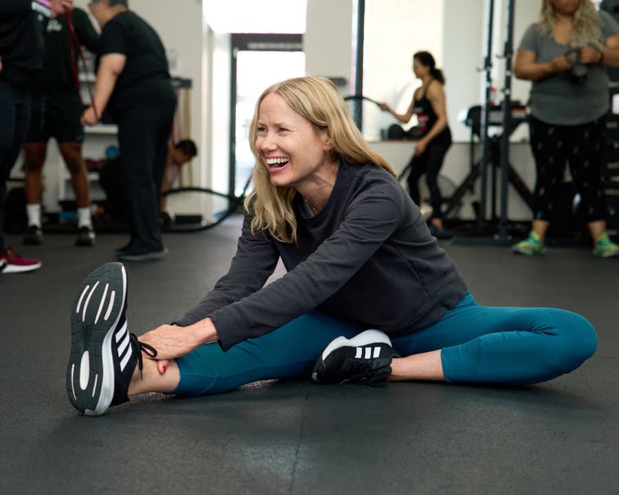 A member sits on the studio floor stretching toward her toes and smiling after a session.